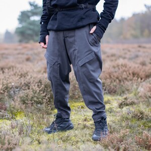 May include: Dark gray cargo pants with large pockets and a relaxed fit. The pants are paired with black athletic shoes and a black jacket. The person is standing in a field of brown and green vegetation.