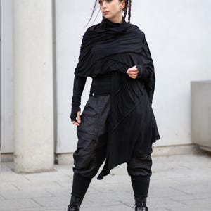 K&ouml;nnte beinhalten: Eine Frau in einem schwarzen Outfit. Das Ensemble umfasst ein lang&auml;rmliges Oberteil, einen geschichteten Schal, eine Cargohose und Kampfstiefel. Das Outfit ist ganz in Schwarz gehalten und schafft einen stimmigen und stilvollen Look. Der Hintergrund ist eine neutrale wei&szlig;e Wand.