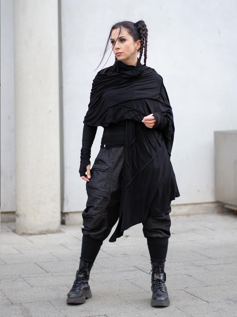 K&ouml;nnte beinhalten: Eine Frau in einem schwarzen Outfit. Das Ensemble umfasst ein lang&auml;rmliges Oberteil, einen geschichteten Schal, eine Cargohose und Kampfstiefel. Das Outfit ist ganz in Schwarz gehalten und schafft einen stimmigen und stilvollen Look. Der Hintergrund ist eine neutrale wei&szlig;e Wand.