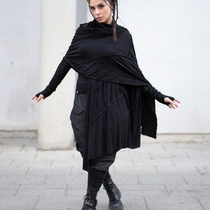 K&ouml;nnte beinhalten: Eine Frau in einem schwarzen Outfit mit einem drapierten Schal, einem lang&auml;rmeligen Oberteil und einem Rock mit geometrischem Design. Das Outfit umfasst dunkle Hosen und schwarze Kampfstiefel. Der Hintergrund ist eine wei&szlig;e Wand und ein grauer Fliesenweg.