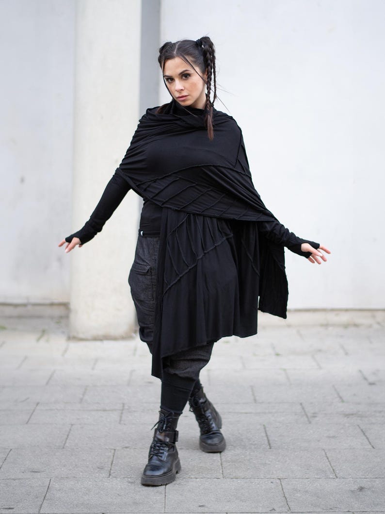 K&ouml;nnte beinhalten: Eine Frau in einem schwarzen Outfit mit einem drapierten Schal, einem lang&auml;rmeligen Oberteil und einem Rock mit geometrischem Design. Das Outfit umfasst dunkle Hosen und schwarze Kampfstiefel. Der Hintergrund ist eine wei&szlig;e Wand und ein grauer Fliesenweg.