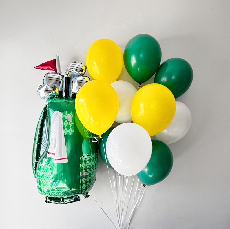 Puede incluir: Un globo de aluminio con forma de bolsa de golf verde con una bandera roja y palos de golf plateados, acompa&ntilde;ado por un racimo de globos de l&aacute;tex redondos en colores amarillo s&oacute;lido, verde oscuro y blanco, atados con cintas de pl&aacute;stico.