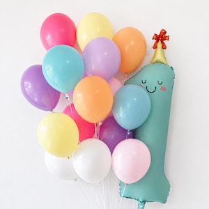 May include: A cluster of colorful latex balloons in shades of pink, yellow, orange, purple, blue, and white, accompanied by a large, teal-colored foil balloon shaped like the number one, featuring a smiling face and a small gold party hat.