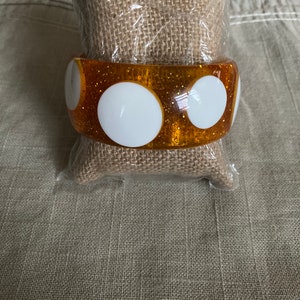 May include: A gold glitter bangle bracelet with white polka dots. The bracelet is made of plastic and has a shiny finish.
