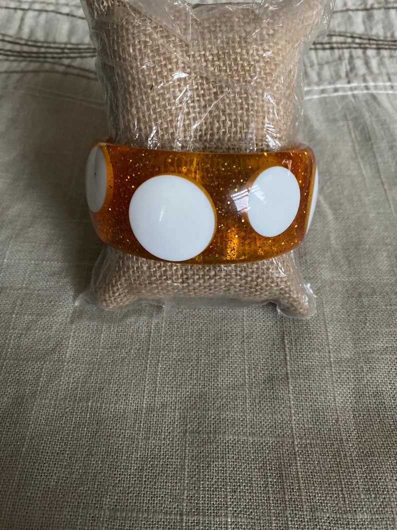 May include: A gold glitter bangle bracelet with white polka dots. The bracelet is made of plastic and has a shiny finish.