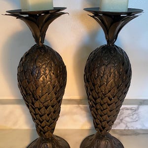 May include: A pair of dark bronze-coloured metal candle holders shaped like pineapples. Each holder features a textured, leaf-patterned body, a circular base, and a flat top plate holding a square, off-white pillar candle.