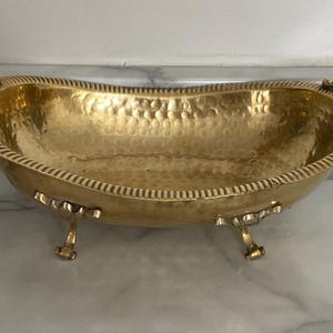 May include: A gold-coloured, hammered metal decorative bowl with ornate handles and feet. The bowl has a textured interior and a decorative rim. The bowl is likely used for display or serving.