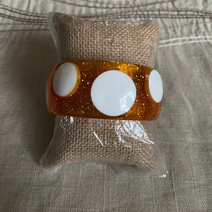 May include: A sparkly orange bangle bracelet with three white circles. The bracelet is on a brown burlap cushion.