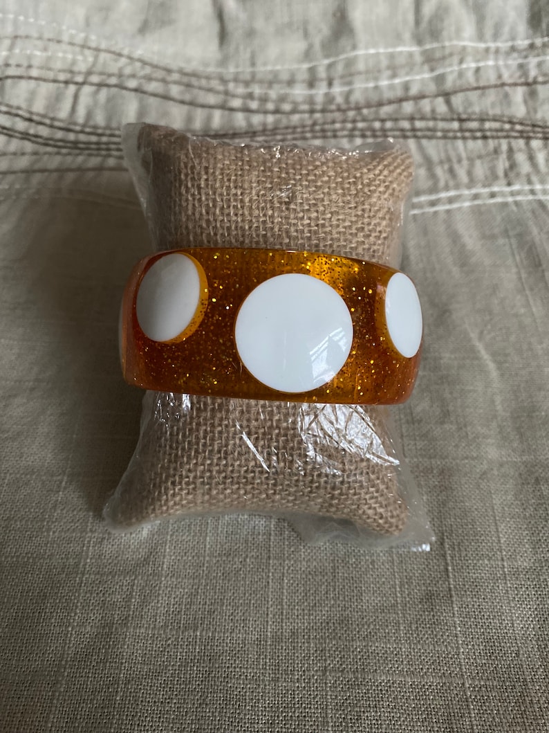 May include: A sparkly orange bangle bracelet with three white circles. The bracelet is on a brown burlap cushion.