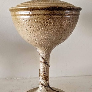 May include: A ceramic lidded jar with a textured, speckled glaze in shades of brown and beige. The jar has a tall, slender base and a rounded lid with a knob on top.