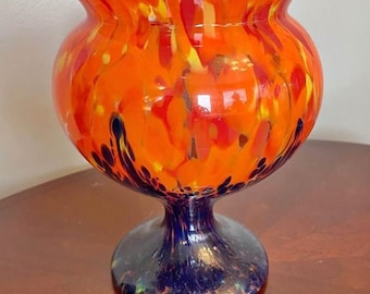May include: A vibrant, hand-blown glass vase with a bulbous orange body and a flared rim. The vase features a speckled pattern of yellow, red, and dark blue. It sits on a dark blue pedestal base, creating a striking contrast.