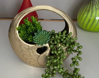 May include: A gold-colored, decorative planter with a handle, filled with green artificial succulents and trailing greenery. The planter has a textured surface and a swan-like design. Two decorative vases are in the background.