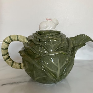 May include: A ceramic teapot shaped like a cabbage, with a bamboo-style handle. The lid features a white bunny figurine with pink ears. The teapot is predominantly green, with cream and tan detailing. A charming kitchenware item.