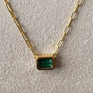May include: A gold-colored necklace with a rectangular emerald-cut green gemstone pendant. The chain is made of small, rectangular links. The necklace is displayed on a textured, light-colored surface.