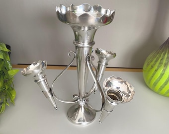 May include: A silver-colored, Art Nouveau-style flower vase. The vase has a central, trumpet-shaped body with a scalloped top and three smaller, cone-shaped flower holders. The design includes curved, decorative supports.