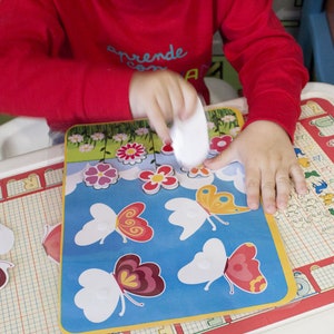 Puede incluir: Las manos de un niño colocando una pegatina blanca sobre una pegatina de mariposa azul y amarilla en una alfombra de aprendizaje colorida. La alfombra tiene un patrón de cuadrícula y presenta letras, números e imágenes de animales. La alfombra está sobre una mesa blanca.