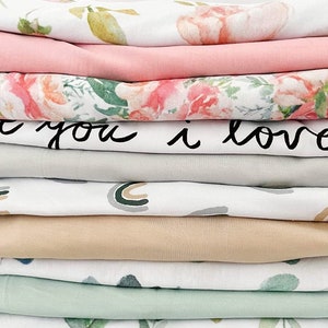 May include: A stack of 10 fabric swaddles in various patterns. The swaddles feature floral prints, a rainbow design, and a solid beige color. The top swaddle has the text "to you i love" written in black.