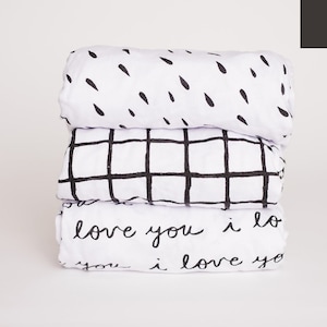 May include: A set of three white fitted crib sheets with black geometric patterns. The sheets feature a black and white raindrop pattern, a black and white grid pattern, and a black and white script pattern that says "love you i love you".