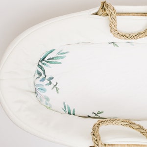 May include: A white oval baby bassinet with a woven rope handle. The interior features a white fabric with a watercolor botanical print in shades of green and blue. The bassinet is designed for newborns.