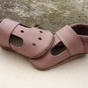 May include: A pair of soft, dusty rose-colored baby shoes. The shoes have a T-strap design with small square cutouts and a strap closure. The soles are a light brown color, and the interior is lined with a darker pink suede-like material.