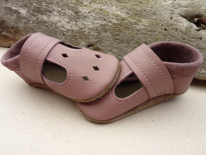 May include: A pair of soft, dusty rose-colored baby shoes. The shoes have a T-strap design with small square cutouts and a strap closure. The soles are a light brown color, and the interior is lined with a darker pink suede-like material.