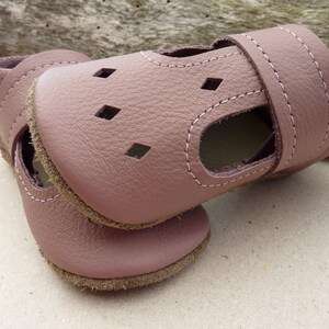 May include: A pair of soft, mauve-colored baby sandals. The sandals are made of leather with a stitched design and diamond-shaped cutouts. The soles are a contrasting brown color, and the ankle strap has a hook and loop closure.