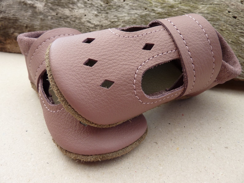 May include: A pair of soft, mauve-colored baby sandals. The sandals are made of leather with a stitched design and diamond-shaped cutouts. The soles are a contrasting brown color, and the ankle strap has a hook and loop closure.