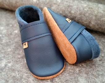 Puede incluir: Un par de zapatos de bebé de cuero azul marino con una suela de ante marrón claro. Los zapatos tienen un emblema en forma de estrella en un pequeño parche de cuero marrón claro. El interior está forrado con una tela azul suave.