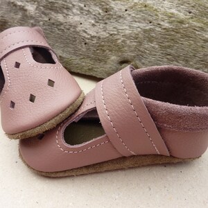 May include: A pair of dusty rose leather baby shoes. The shoes have a strap with a velcro closure and diamond-shaped cutouts. The soles are a light brown color, and the shoes are stitched with white thread.