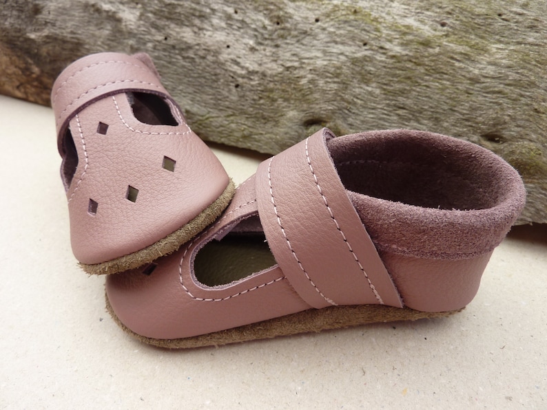 May include: A pair of dusty rose leather baby shoes. The shoes have a strap with a velcro closure and diamond-shaped cutouts. The soles are a light brown color, and the shoes are stitched with white thread.