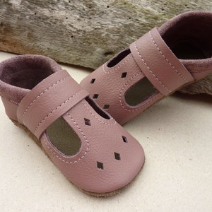 May include: A pair of soft, dusty rose leather baby shoes. The shoes have a T-strap design with diamond-shaped cutouts on the toe. The interior and ankle cuffs are a darker shade of rose. The shoes are designed for comfort and style.