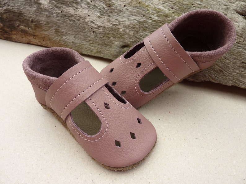 May include: A pair of soft, dusty rose leather baby shoes. The shoes have a T-strap design with diamond-shaped cutouts on the toe. The interior and ankle cuffs are a darker shade of rose. The shoes are designed for comfort and style.