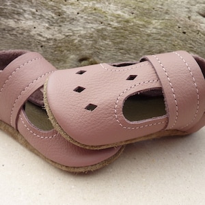 May include: A pair of dusty rose leather baby shoes. The shoes have a strap across the top with white stitching and small diamond-shaped cutouts. The soles are a light brown color, and the interior is a darker rose color.