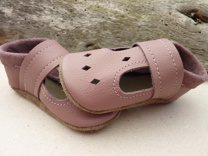 May include: A pair of dusty rose leather baby shoes. The shoes have a strap across the top with white stitching and small diamond-shaped cutouts. The soles are a light brown color, and the interior is a darker rose color.