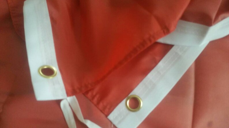 May include: Close-up of a red fabric flag with white trim and two gold grommets. The flag is folded in half, showing the seam and the white trim.
