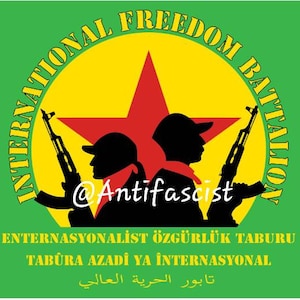 May include: A green flag with a yellow circle containing a red star and two black silhouettes of people holding rifles. The text "INTERNATIONAL FREEDOM BATTALION" is written around the circle in yellow. The text "ENTERNASYONALİST ÖZGÜRLÜK TABURU TABŪRA AZADÎ YA ÎNTERNASYONAL تابور الحرية العالي" is written below the circle in yellow.