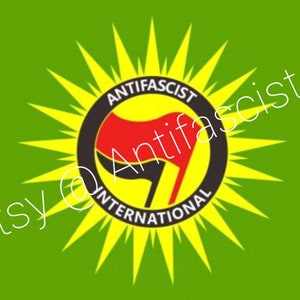 May include: A green flag featuring a yellow sunburst design. The sunburst surrounds a black circle containing a red and black fist symbol. The text "Anti-Fascist International" is inscribed around the circle.