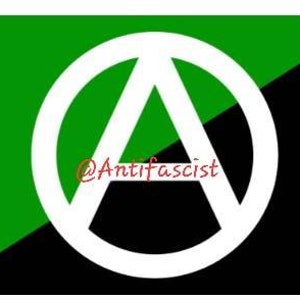 May include: A black and green flag with a white circle containing a stylized letter "A" in the center. The text "@Antifascist" is written in red below the circle.