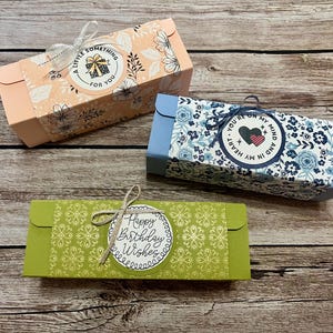 May include: Three rectangular gift boxes with decorative floral patterns and text. One is peach with a floral design and the text "A LITTLE SOMETHING FOR YOU". Another is blue with a floral pattern and the text "YOU'RE ON MY HEART AND IN MY MIND". The third is green with the text "Happy Birthday Wishes".