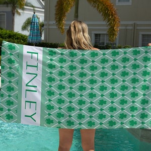 May include: A green beach towel with a white shamrock pattern and the name "FINLEY" printed on the side. The towel is being held by a person standing by a swimming pool.