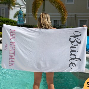May include: White beach towel with the text "Bride" in black cursive and "Mrs. Whitman 2022" in pink lettering.