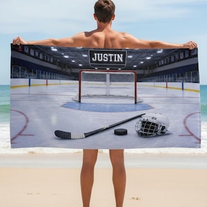 May include: A rectangular beach towel featuring a hockey rink scene with the name "JUSTIN" above. The towel displays a hockey net, stick, puck, and helmet. The background shows a hockey arena.