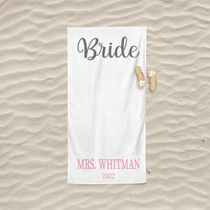 May include: White beach towel with the word "Bride" in black cursive lettering and the text "MRS. WHITMAN 2022" in pink lettering.
