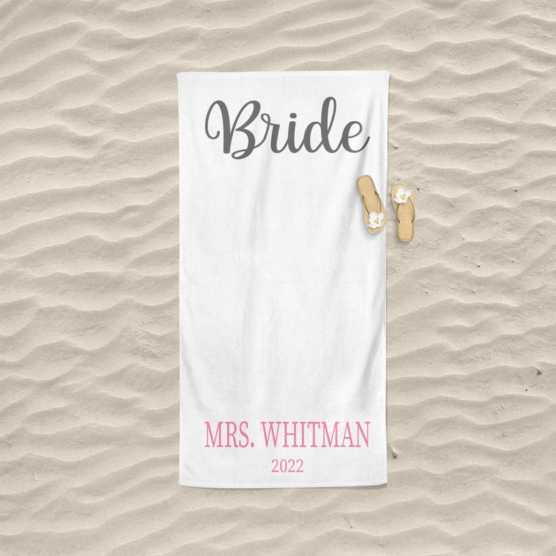 May include: White beach towel with the word "Bride" in black cursive lettering and the text "MRS. WHITMAN 2022" in pink lettering.