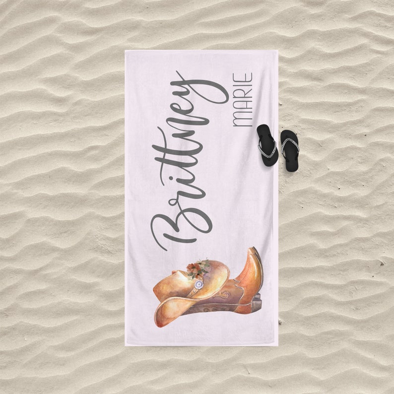May include: White beach towel with the name "Brittany Marie" written in gray cursive script. A watercolor illustration of a brown cowboy hat and boot is in the bottom right corner.