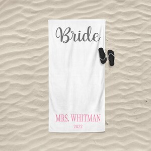 May include: White beach towel with the word "Bride" in gray cursive lettering and the text "MRS. WHITMAN 2022" in pink lettering. A pair of black flip flops are on the towel.