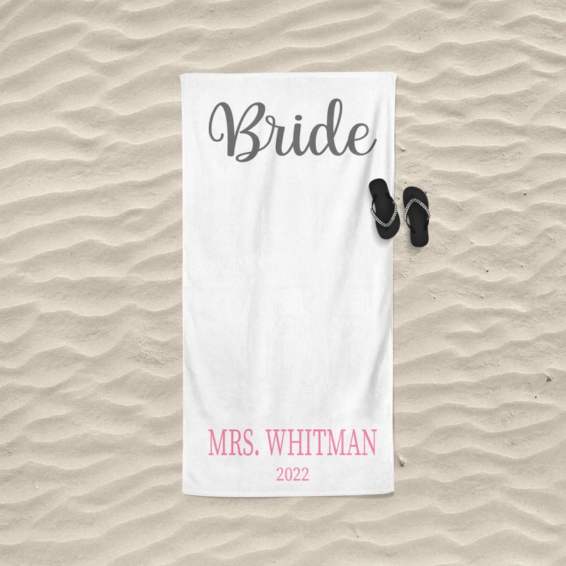 May include: White beach towel with the word "Bride" in gray cursive lettering and the text "MRS. WHITMAN 2022" in pink lettering. A pair of black flip flops are on the towel.