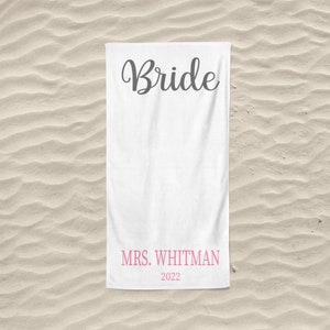 May include: White beach towel with the word "Bride" in black cursive lettering and the text "MRS. WHITMAN 2022" in pink lettering.