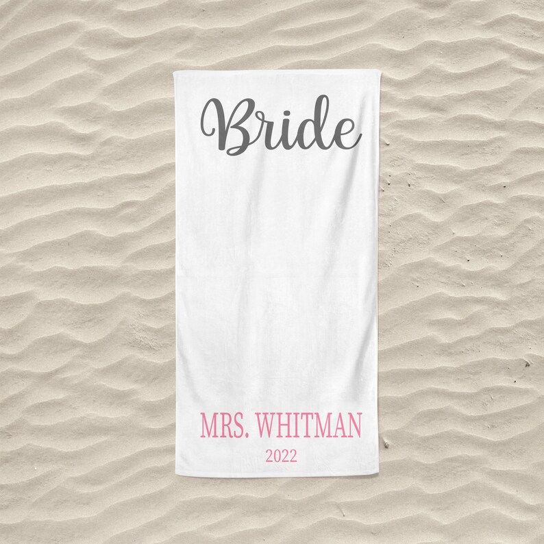 May include: White beach towel with the word "Bride" in black cursive lettering and the text "MRS. WHITMAN 2022" in pink lettering.