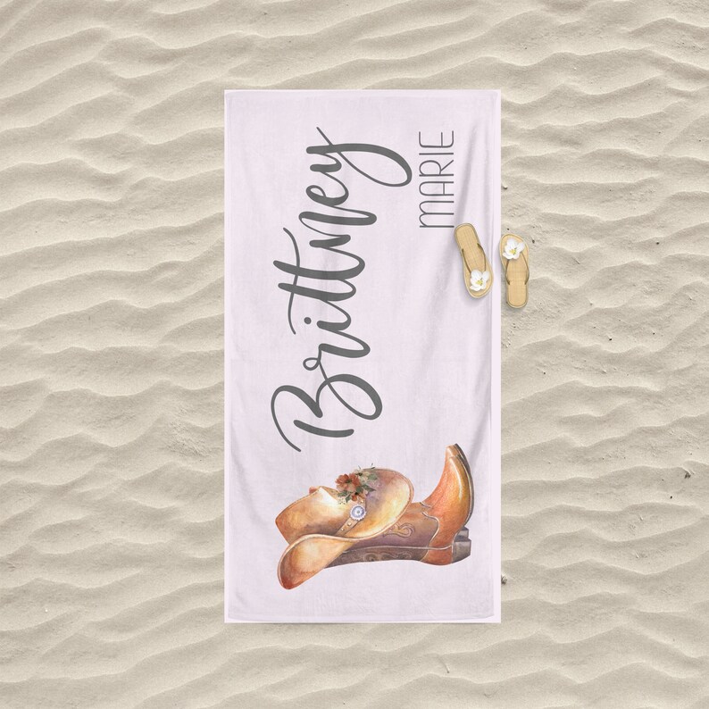 May include: White beach towel with the name "Brittany Marie" written in black cursive script. A watercolor illustration of a brown cowboy boot and hat with floral accents is in the bottom center of the towel.